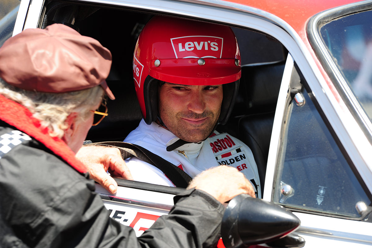 Motor racing driver Peter Brock wearing a red helmet sits in the front seat of a car