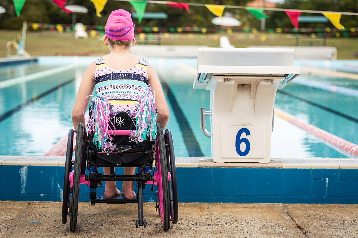 A girl in a wheelchair wearing a swimming costume and swim cap waits next to the starting blockat the pool