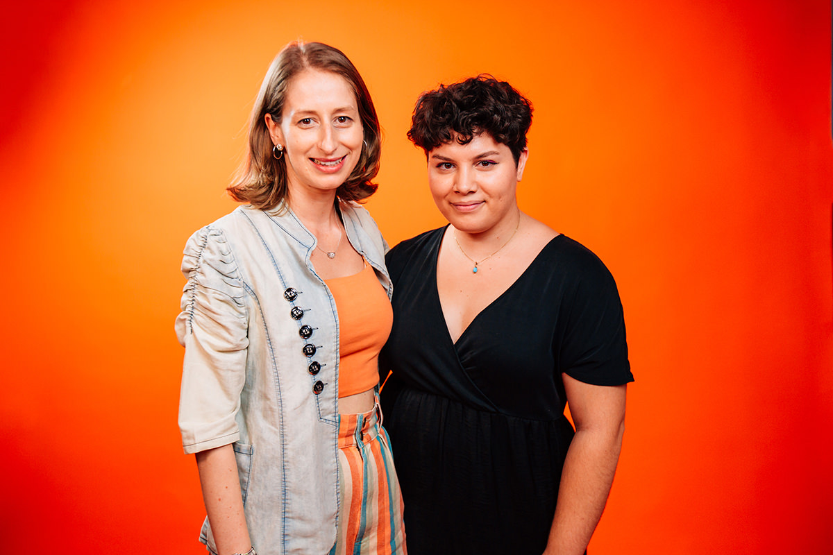 Headshot of Hayley Adams and Michelle Melky, standing together in front of orange background.