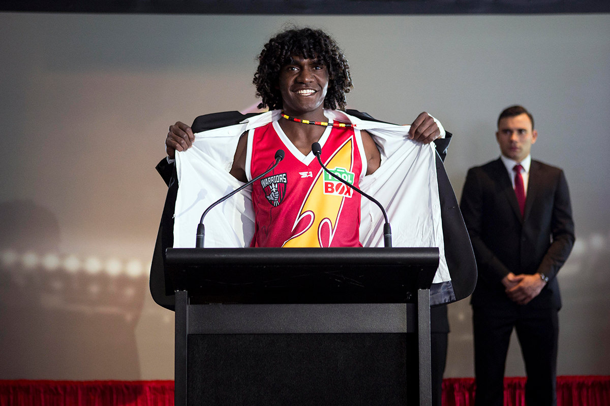 A man stands at a lectern, he has his jacket pulled open showing his AFL jersey 