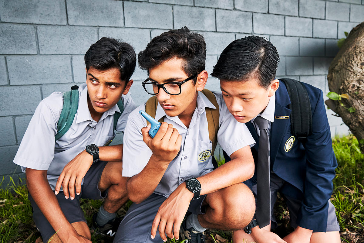 A group of three teenage boys wearing school uniform huddle around a walkie talkie