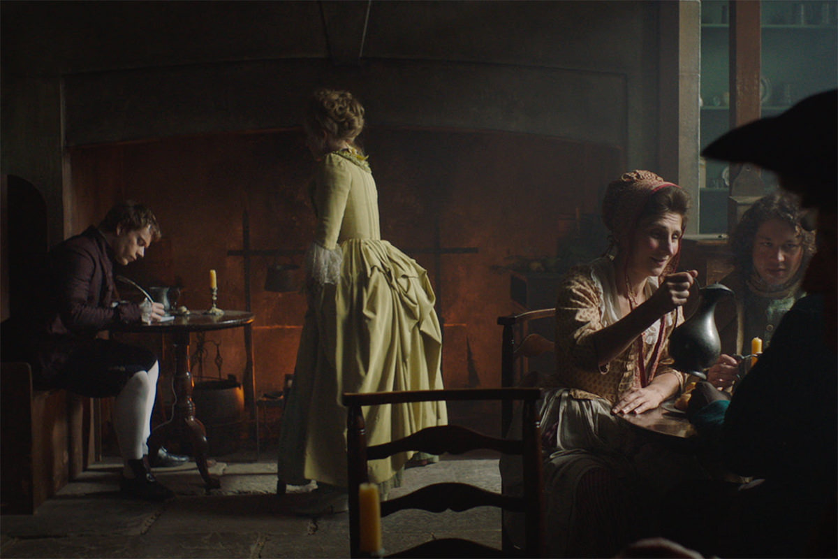 A scene from Harlots, a man sits at a table in front of a fire writing with a quill. Nearby, a group of men and women share a drink