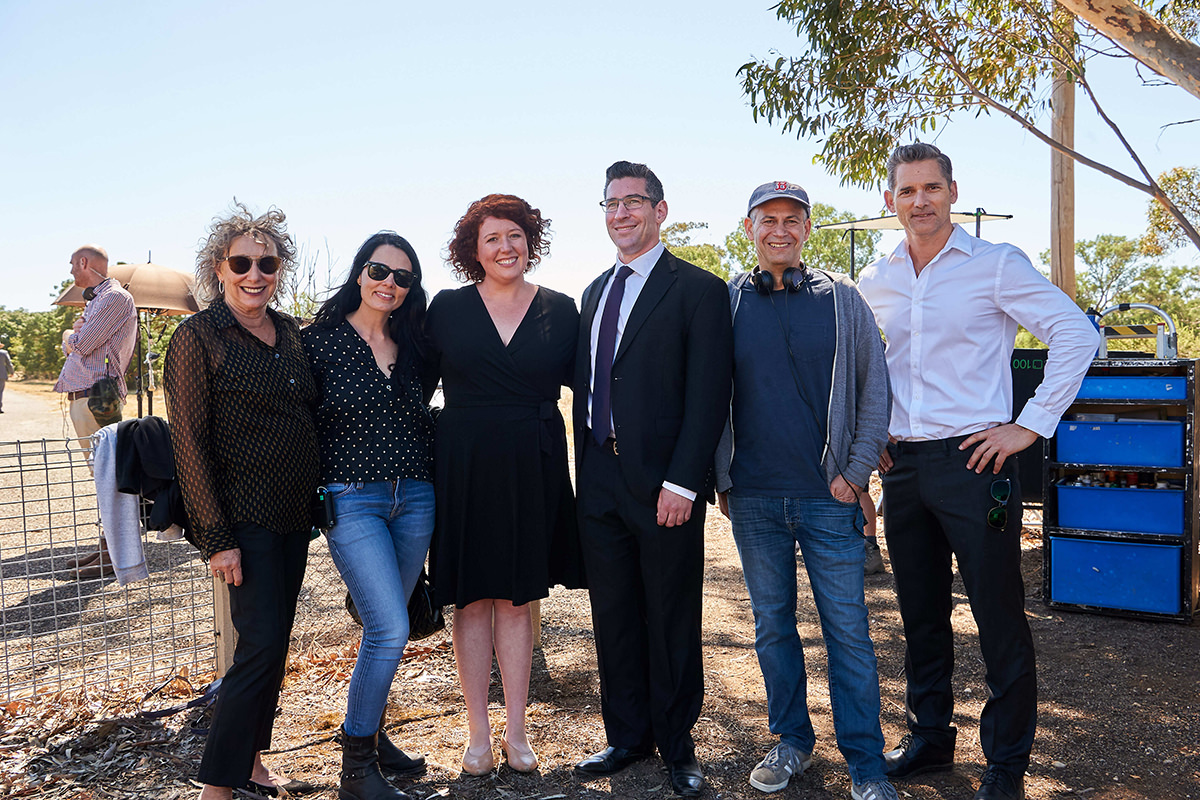 A group of six people stand together smiling at the camera. Behind them is a film crew. They are in the Australian country.