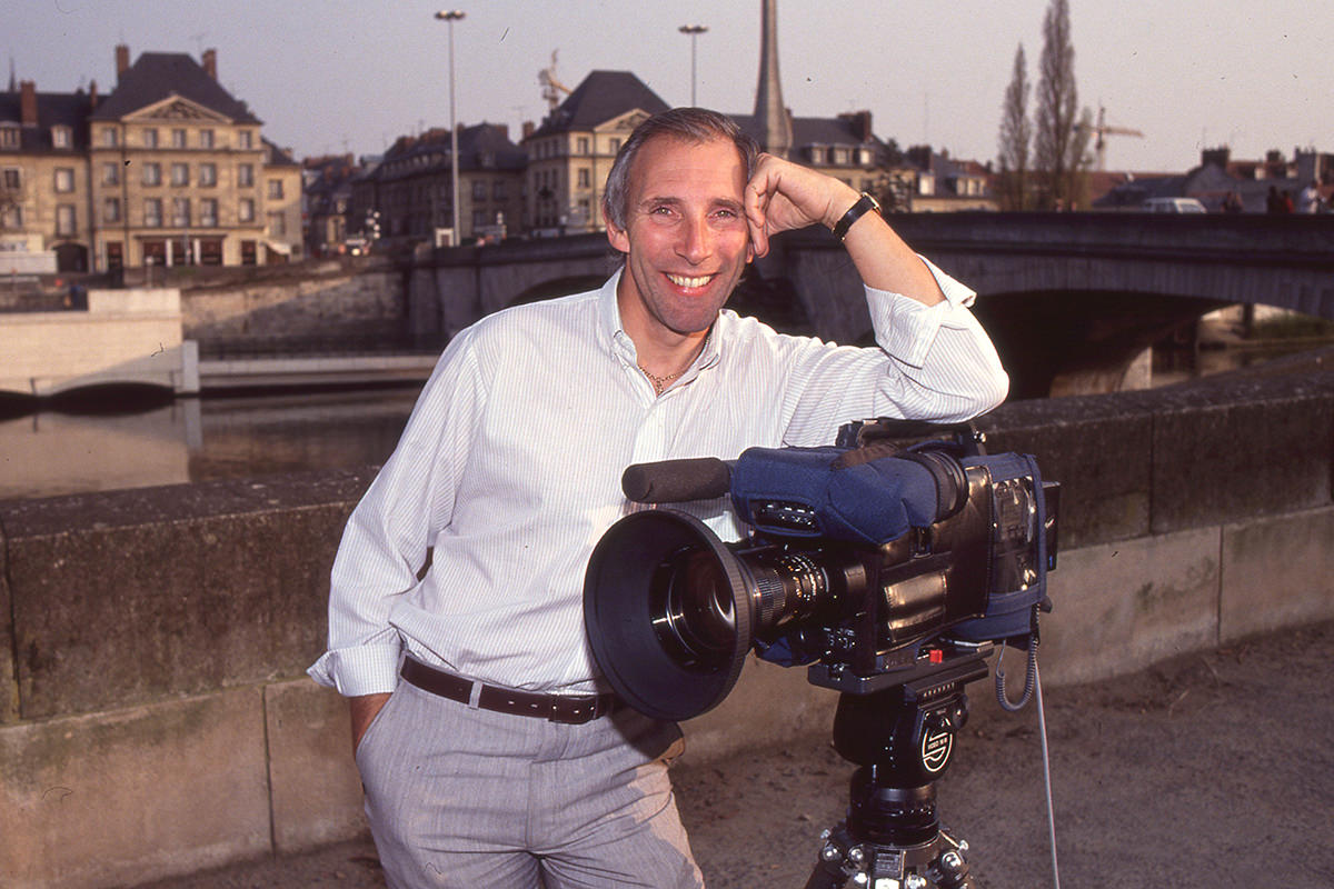 Sports broadcaster Phil Liggett leans against a video camera, smiling