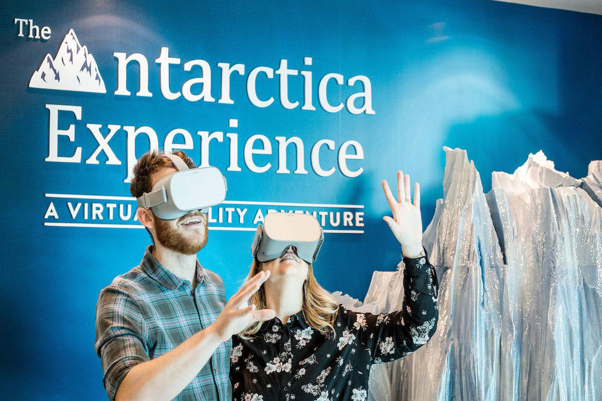 Two people wearing VR headsets stand in front of a sign that says 'The Antarctica Experience'.
