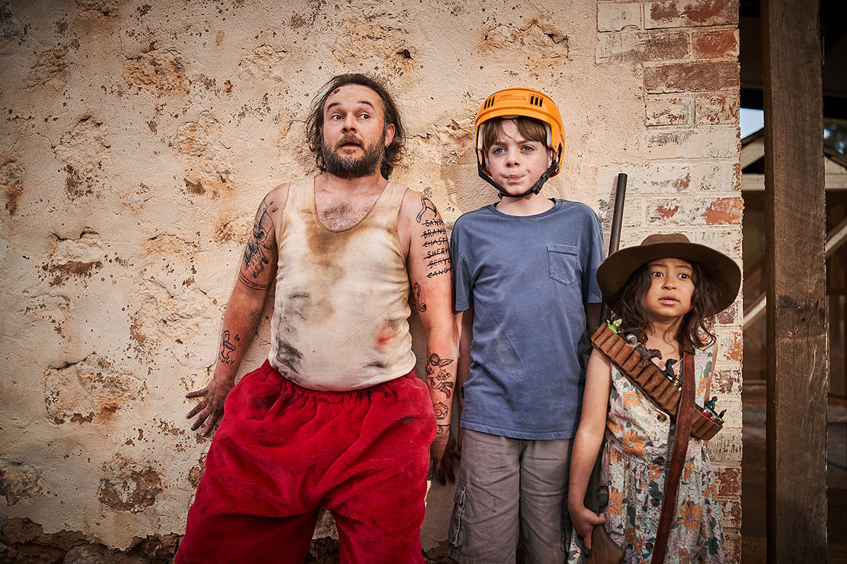 A man dressed in red Santa pants and two children stand with their backs against a wall