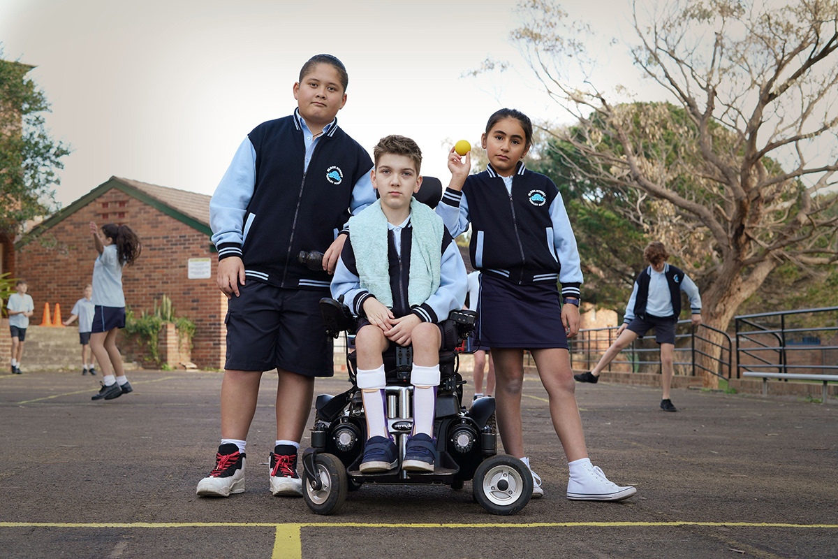 Three children huddle together on a handball court in the school playground. One is holding a handball, another is in a wheelchair