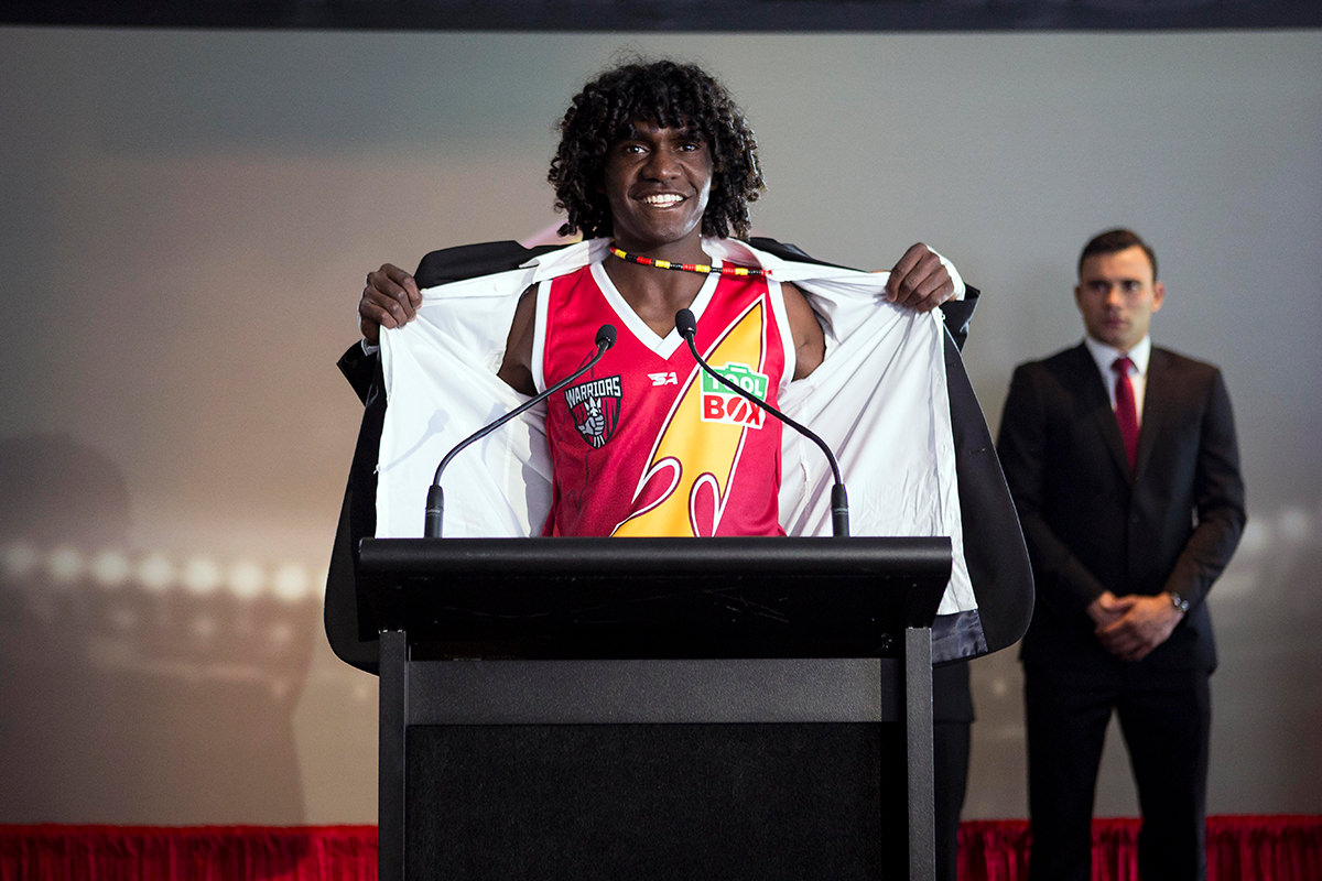 A man stands at a lectern, he has his jacket pulled open showing his AFL jersey