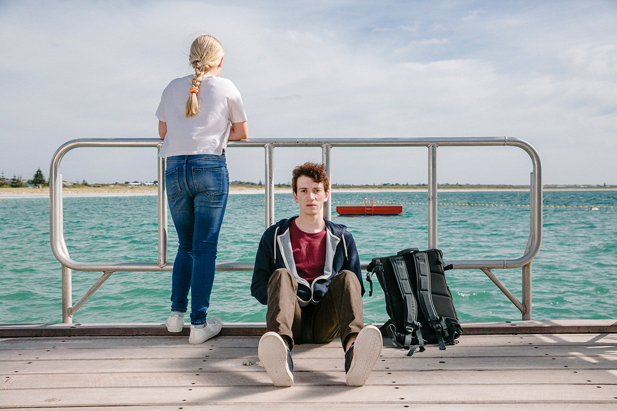 Two teenagers on a wharf, overlooking the water. One is sitting down, the other is looking out at the view