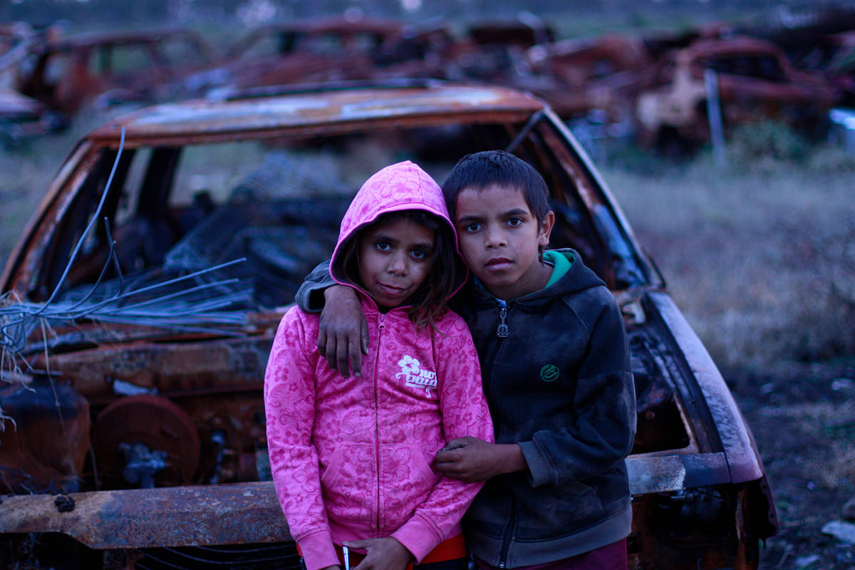 Two children stand in front of a burnt out car. One has their arm around the other.