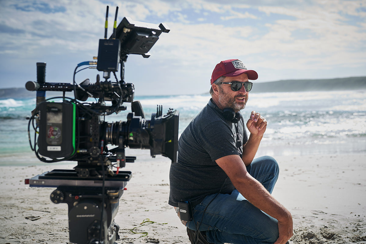 Director Gregor Jordan is kneeling down on the beach. A large film camera is to his left