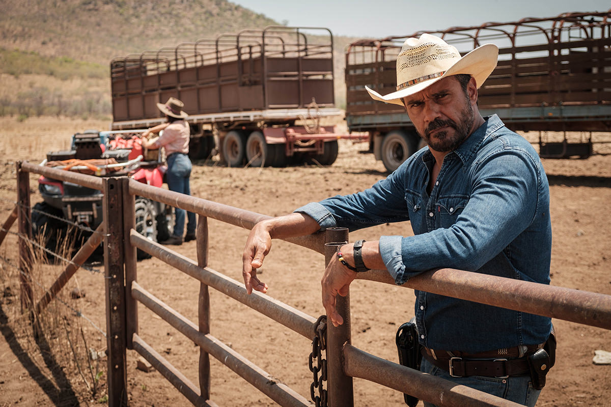 Detective Jay Swan stands in a remote landscape, his hands resting on a fence